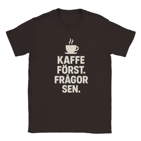 Mörkbrun t-shirt med vit text: "Kaffe först. Frågor sen." och en enkel illustration av en rykande kaffekopp ovanför.