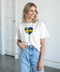 Svensk flagga hjärta på vit T-shirt, modell med blont hår och jeans.