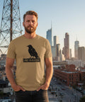 Svart kråka på skylt "ADAPT OR DISAPPEAR" på beige ekologisk T-shirt, man i stadsmiljö.