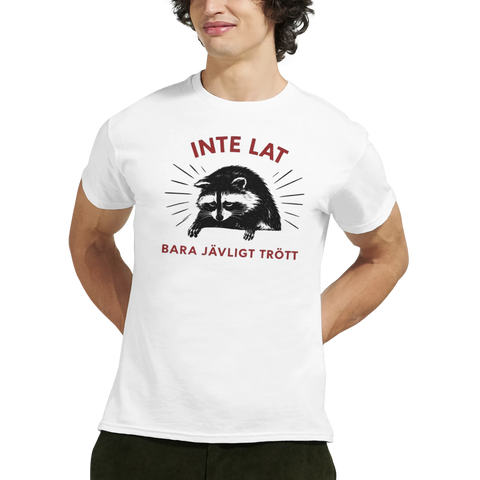 Sarkastisk tvättbjörn med text "INTE LAT BARA JÄVLIGT TRÖTT" på vit t-shirt.