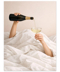 Fotografi med fokus på händer som håller en champagneflaska och glas i en vit obäddad säng.