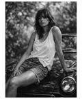 Svartvitt fotografi av en kvinna i linne och jeansshorts som sitter på motorhuven av en klassisk bil i naturen.