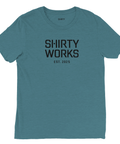 Shirty Works logga, EST. 2025, i svart på mörk turkos triblend t-shirt.