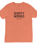 Shirty Works EST. 2025" svart text på persikofärgad triblend t-shirt.