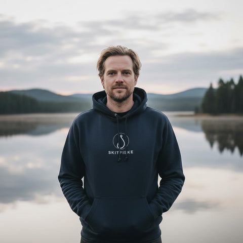 Man i Skitfiske-hook hoodie vid spegelblank sjölandskap.