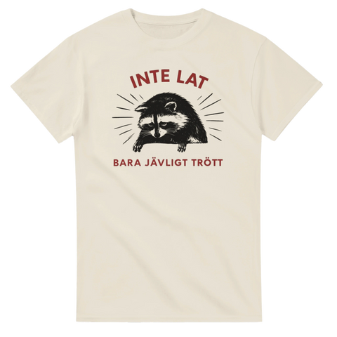 Trött tvättbjörn med text "INTE LAT BARA JÄVLIGT TRÖTT" på gräddvit t-shirt.