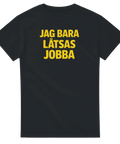 Jag bara låtsas jobba" texttryck i gul, fet stil på en svart t-shirt.