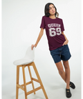 Vit text "QUEEN 69" med dubbel kontur på vinröd t-shirt, sportig college-stil.