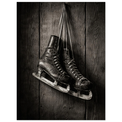 Svartvita slitna läderskridskor med snörning som hänger i sina skosnören mot en rustik och väderbiten trävägg.