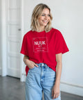 Nuuk periodiskt system-design på röd t-shirt, bärs av leende kvinna i ljusa jeans.