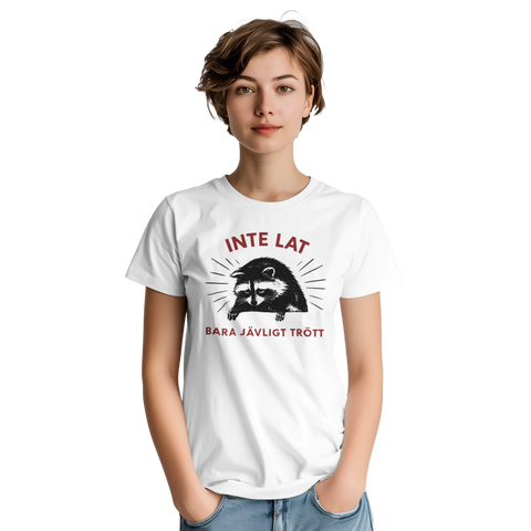 Sömnig tvättbjörn med text "INTE LAT BARA JÄVLIGT TRÖTT" på vit t-shirt.