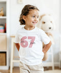 Rosa "67" med texten "Six + Seven" på vit barnt-shirt.