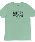 Svart "SHIRTY WORKS EST. 2025" tryck på ljusgrön triblend t-shirt.